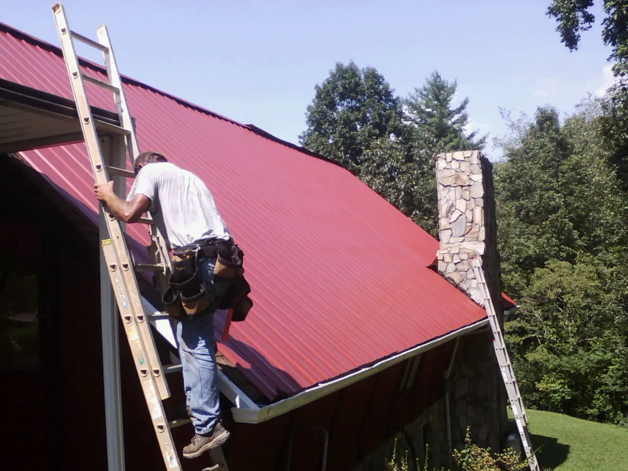 Roofer on a ladder beside a red metal roof and stone chimney — Soffit & Fascia Repair by Roofing Brothers in Goochland